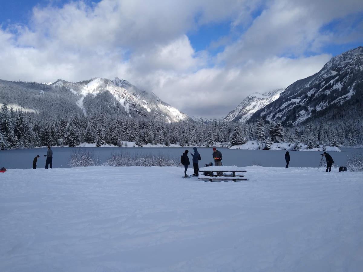 A frozen lake with an open flat area of snow in front with several groups of people, and snow covered mountains in the distance.