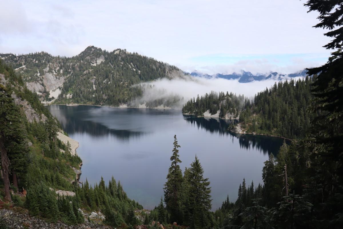 A deep blue mountain lake, surrounded by forested mountainsides, with a misty cloud rolling onto the surface through the distant trees.