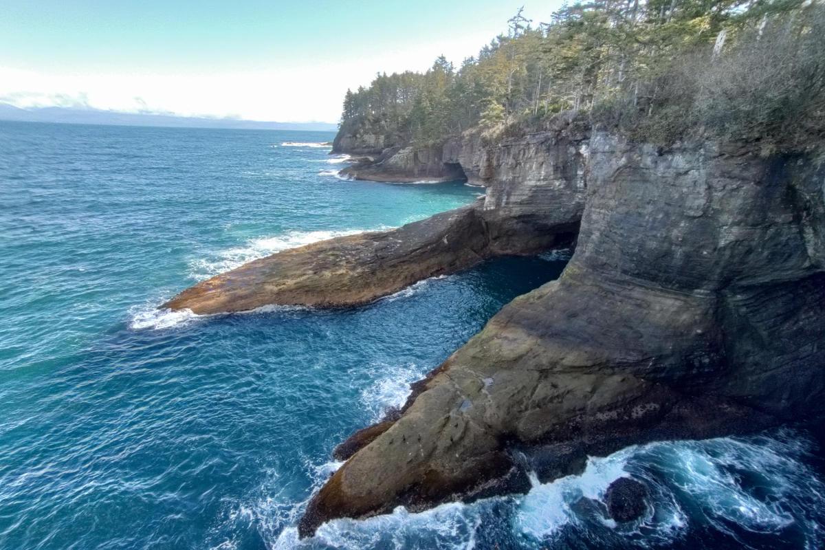 Several arms of hard worn rock thrust from a steep cliff face out into a tumultuous sea.