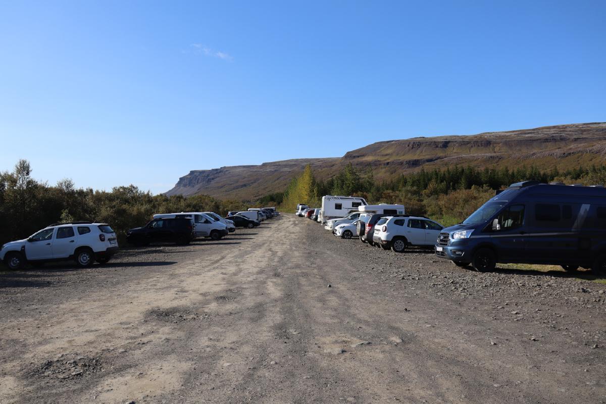 Photo of a gravel car park, with cars on either side, and trees in the distance.