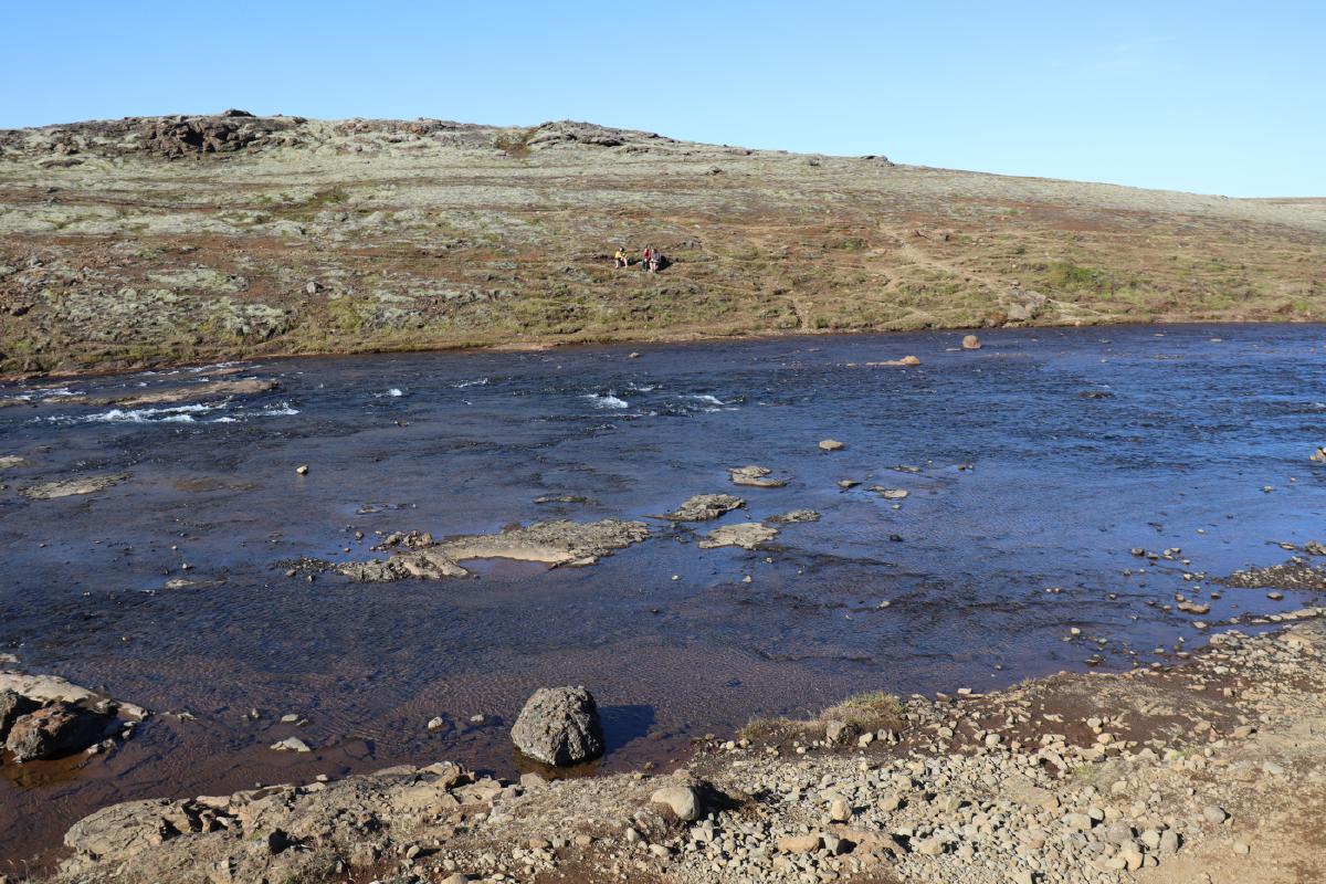 Photo of a fairly calm and gentle, but wide river, with a rocky surface and grass hill on the other side.