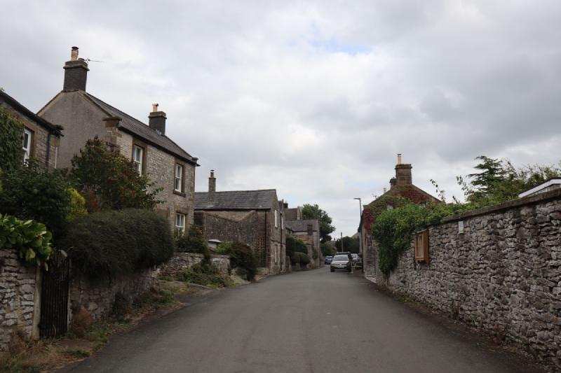 The main road through the village. Stone buildings on the left, a stone wall on the right, and no pavement.