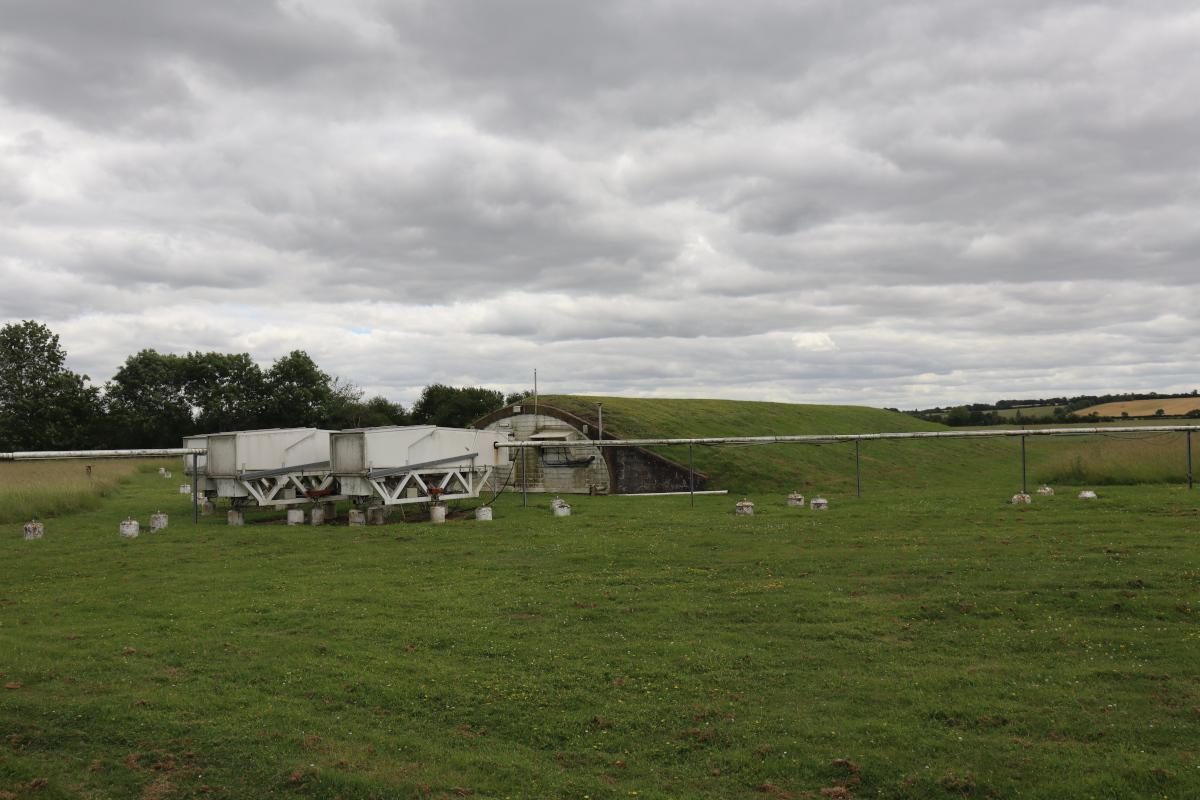 A bunker covered in grass with some white boxes out front, connected to the bunker by white pipes.