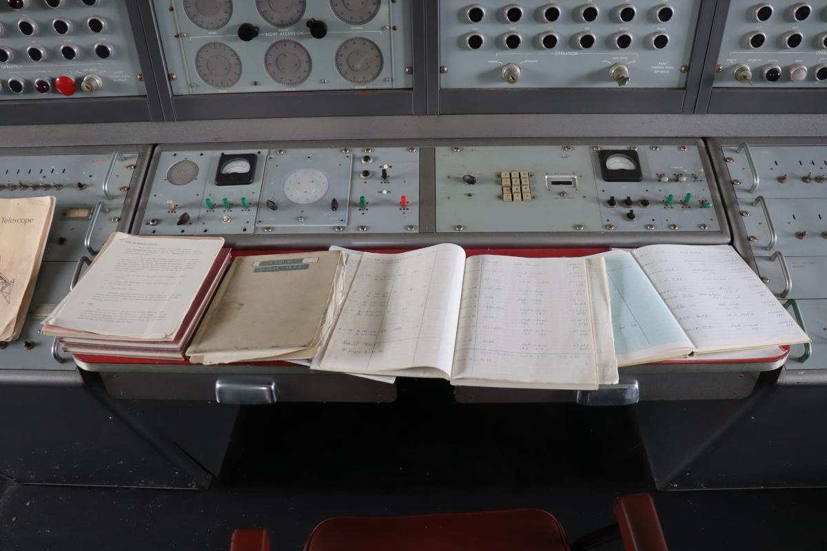 A control panel, four units wide, with various dials and buttons, and a desk covered with log books.