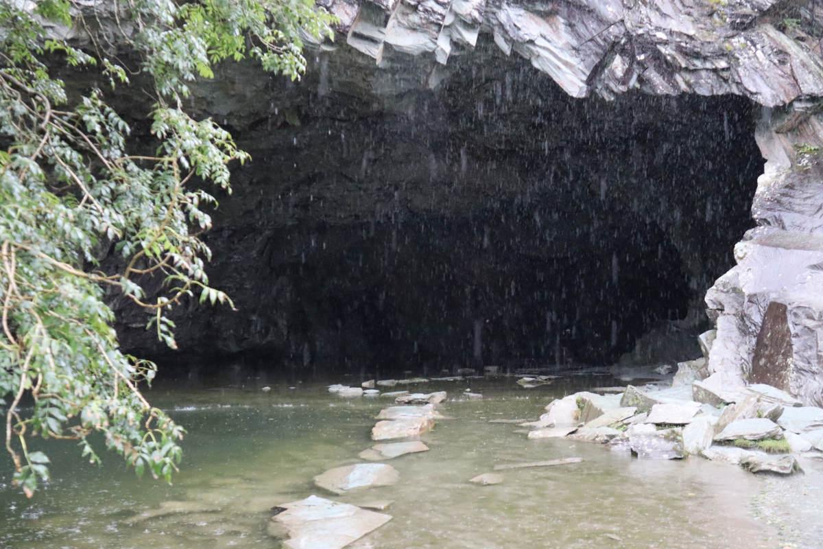 A dark cave entrance, filled with a pool of water, with slabs of stepping stones leading inwards.